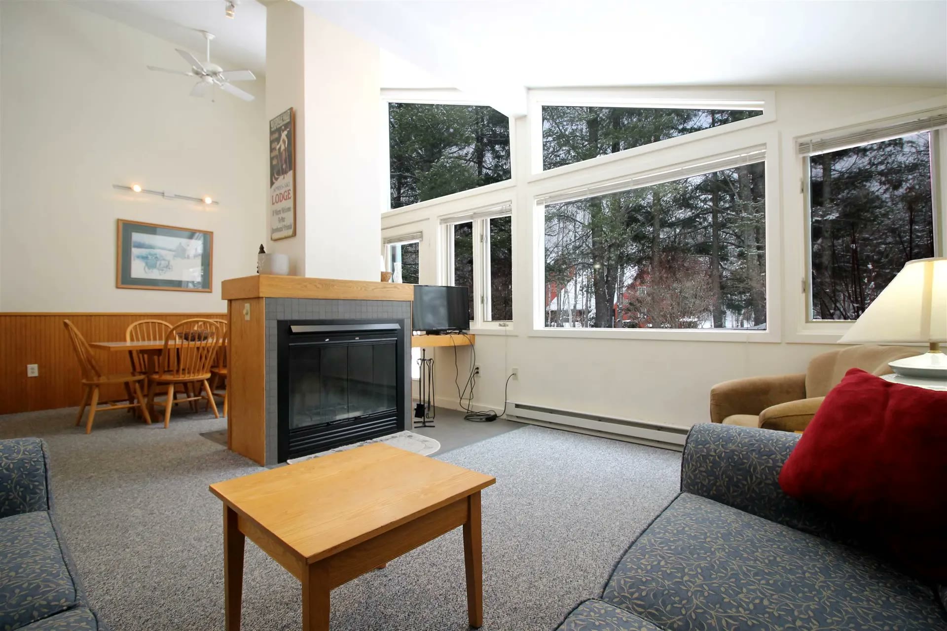 Bright living room with a two-sided fireplace, dining table, and tall windows onto the pines